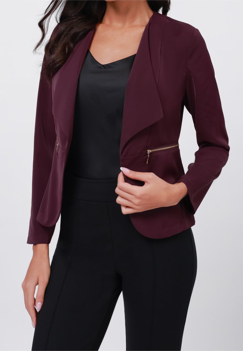 Femme portant un blazer violet foncé avec des détails de fermeture éclair dorée sur un haut en satin noir et un pantalon noir, raccourci au niveau du cou et des cuisses.