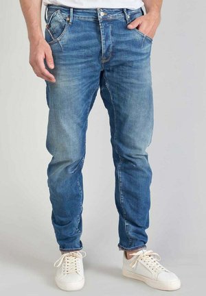 Jean en denim bleu avec une coupe slim, un léger effet usé, un subtil délavage et des détails de couture visibles, portés avec des baskets blanches.