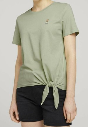 Kobieta nosząca jasnozieloną bluzkę z krótkim rękawem z węzłem z boku i napisem „słońce. słońce. zabawa.”, połączoną z czarnymi szortami.