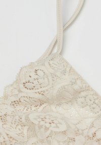 Bralette en dentelle beige avec des motifs floraux et des détails raffinés, doté de bretelles ajustables et de bords délicats. Texture douce et design léger.