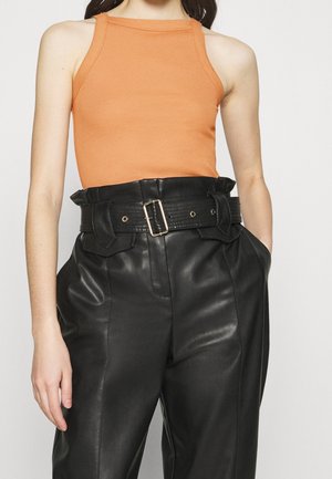 Oranges gerippter Tanktop kombiniert mit hoch taillierten schwarzen Lederhosen, die einen breiten Gürtel und eine große Schnalle aufweisen und eine strukturierte Silhouette erzeugen.