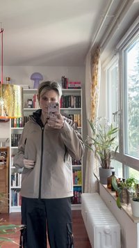 Ljusbeige zip-up jacka med huva, med en svart dragkedja och elastiska cuffs, bärs över svarta cargobyxor, i en väl upplyst inomhusmiljö.