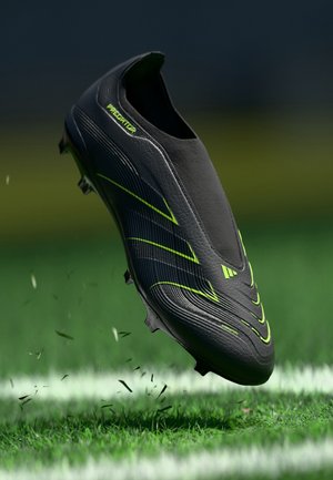 Chaussure de football Adidas Predator noire et vert néon flottant au-dessus de l'herbe avec de petits morceaux d'herbe volant autour.