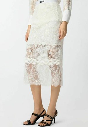 Donna che indossa una gonna midi in pizzo bianco con motivo floreale e sandali neri con cinturini e tacchi alti, in piedi davanti a uno sfondo semplice.