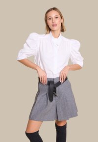 Hvit skjorte med puffermer, grå plissert skjørt med svart belte, knelang mørk sokker, glatt stoff og strukturert design. Nøytral bakgrunn.