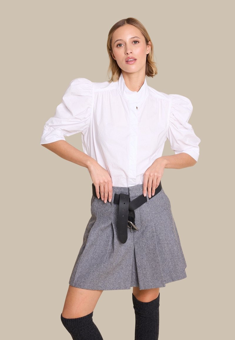 Hvit skjorte med puffermer, grå plissert skjørt med svart belte, knelang mørk sokker, glatt stoff og strukturert design. Nøytral bakgrunn.