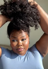 Lockigt svart hår uppsatt, iförd en ljusblå, figurformad topp. Mjukt ljus framhäver hudton och textur, med synliga arm- och ansiktsdrag.