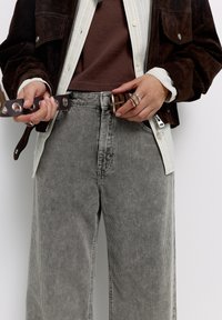 Bruine leren jas over een bruine blouse, grijze corduroy broek met brede pijpen en verticale strepen, houdend een bruine riem met metalen accenten.