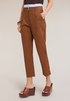 Frau trägt braune, hoch taillierte, knöchellange Hosen mit Falten und großen Seitentaschen, kombiniert mit braunen Keilsandalen und Armbändern.