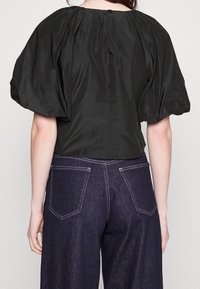 Blusa nera a maniche corte con scollatura arricciata e chiusura a bottone nella parte posteriore, abbinata a jeans a gamba larga blu scuro con cuciture bianche.