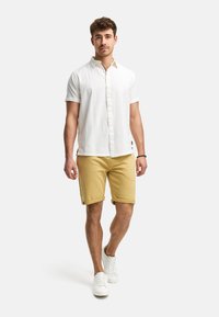 Camisa blanca de manga corta con botones y un patrón texturizado, combinada con pantalones cortos amarillos. Las zapatillas son blancas. Conjunto de verano simple y casual.