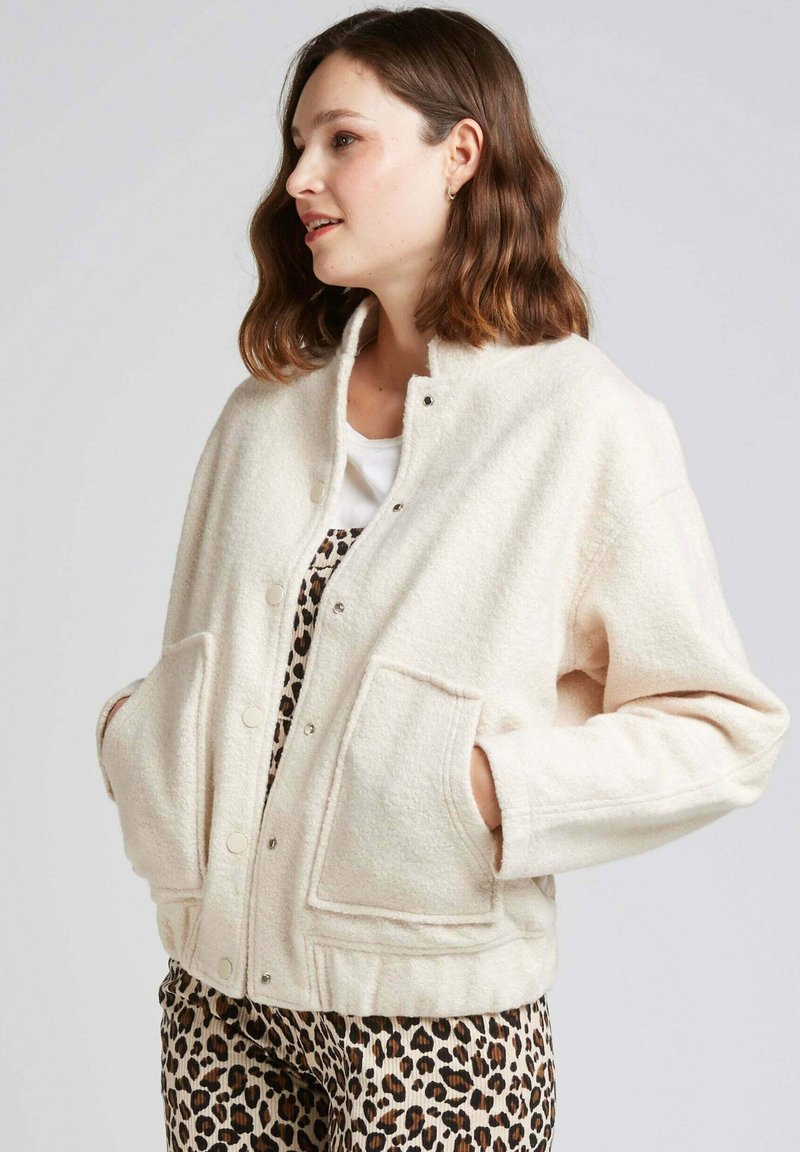 Veste en laine couleur crème avec un col arrondi, des boutons-pression et des poches en patch. Présente une finition texturée. Portée par-dessus une tenue à motif léopard.