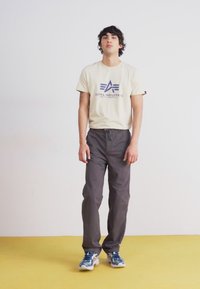Krem t-skjorte med marineblå logo, kombinert med grå bukser og blå sneakers. Står på gul gulv mot en hvit bakgrunn.