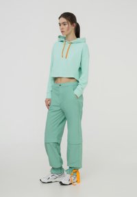 Hoodie cropped verde menta con lacci arancioni abbinato a pantaloni verde menta coordinati, dotati di polsini regolabili e tasche laterali. Sneakers bianche.