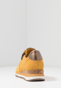 Zapato de ante amarillo con un acento de bronce en el talón, plantilla de corcho texturizada y panel lateral texturizado. Presenta una suela de goma blanca con agarre.