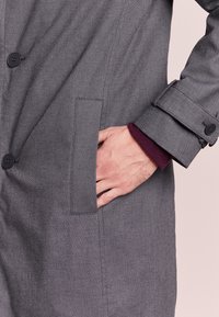 Szary płaszcz zapinany na guziki, wykonany z teksturowanej tkaniny, z boczną kieszenią oraz kontrastującym, bordowym mankietem. Guziki są czarne i wyraźnie widoczne.