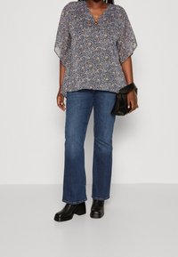 Blusa floreale blu con maniche ampie, scollatura a V e dettaglio in anello bronzeo. Abbinata a jeans blu a gamba dritta e stivaletti neri con tacco bloccato.