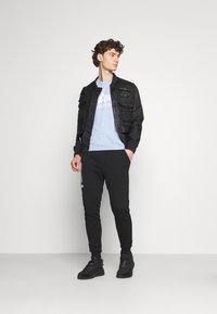 Černá bomber bunda s několika předními kapsami, světle modré tričko s grafickým designem, černé slim-fit tepláky a černé tenisky.