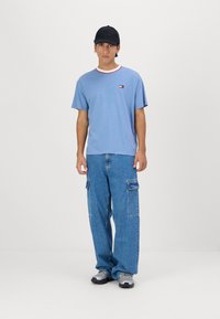 Camiseta de algodón azul claro con un cuello de rayas rojas, pequeño logo en el pecho, combinada con pantalones cargo azules holgados y zapatillas grises.