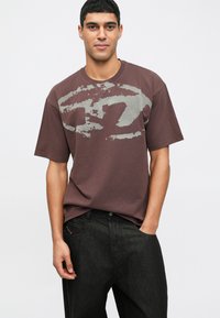 Camiseta castanha com gola redonda, apresentando uma grande impressão gráfica abstrata cinza na frente. Combinada com jeans escuros.