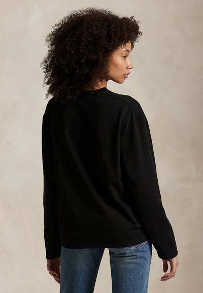 Kobieta z kręconymi włosami, ubrana w czarną koszulkę z długim rękawem i niebieskie dżinsy, stojąca bokiem na jednolitym tle.