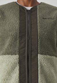 Veste en polaire verte avec un panneau texturé contrastant, dotée d'une fermeture zippée centrale, d'un col rond et d'un détail de poche horizontale.