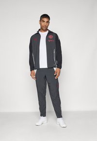 Tuta nera e grigia con accenti rossi, composta da una giacca con zip e pantaloni, realizzata in tessuto morbido. Sneakers bianche completano il look.