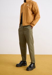 Brun bomberjacka, grön byxor och svarta snörskor. Jackan har logotyp och ribbade ärmar; byxorna har logotyp och rak passform. Texturerad golv.