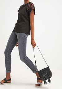 Top nero con maniche a taglio texturizzato, jeans aderenti grigi chiari e borsa nera con frange. Calzature sono sandali neri con una fascia argentata.