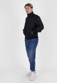 Giacca bomber nera con colletto alto, chiusura a zip e tasche laterali, abbinata a jeans in denim blu e sneakers bianche.