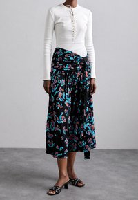 Witte lange mouw top met decoratieve knopen, gecombineerd met een zwarte wikkelrok met een kleurrijk bloemenpatroon. Zwarte slidesandalen met studs.