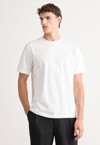 T-shirt en coton blanc à manches courtes, avec un col rond et un détail de logo discret à l'avant. Texture lisse, coupe classique.