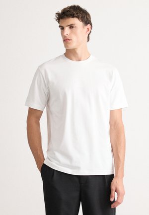 T-shirt en coton blanc à manches courtes, avec un col rond et un détail de logo discret à l'avant. Texture lisse, coupe classique.