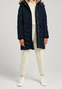 Granatowa kurtka puchowa z kapturem wykończonym sztucznym futrem, z poziomym pikowaniem, zamkiem błyskawicznym i przednimi kieszeniami. Nosi się ją na kremowym swetrze i jasnych spodniach.