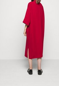 Robe rouge oversize avec manches courtes et fentes latérales. Fabriquée en tissu doux, avec une coupe ample et un design minimaliste. Chaussures noires visibles.
