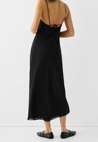 Czarna sukienka midi w kropki z ramiączkami spaghetti, z niskim plecami i wiązanym detalem, zestawiona z płaskimi czarnymi butami na neutralnym tle.
