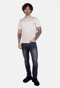 T-shirt bianco a maniche corte con testo stampato, abbinato a jeans scuri slim-fit e sneakers nere. Il modello si trova in piedi con le mani sui fianchi.