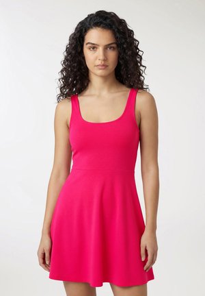 Donna con capelli ricci e scuri che indossa un vestito senza maniche color rosa acceso con scollo a barchetta, in piedi davanti a uno sfondo chiaro e uniforme.