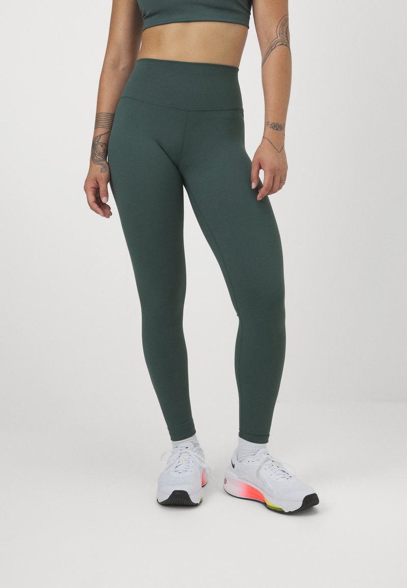 Leggings a vita alta di colore verde scuro, realizzati in un materiale elasticizzato, abbinati a scarpe da ginnastica bianche con accenti rosa e suola nera.