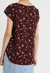 Burgunderbluse mit kurzen Ärmeln, weißem und blauem Blumenmuster, Knopfverschluss hinten, asymmetrischer Saum und weichem Stofftexture.