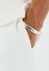 Una pulsera de plata con un logo grabado reposa sobre una muñeca, combinada con una prenda blanca texturizada, mostrando un acabado metálico suave y un diseño simple.