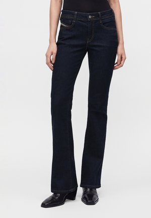 Femme portant un jean bootcut bleu foncé associé à des bottines noires à talons, debout devant un fond clair uni.