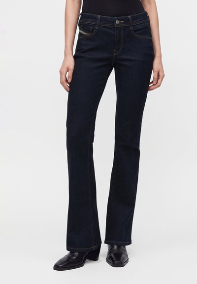 Femme portant un jean bootcut bleu foncé associé à des bottines noires à talons, debout devant un fond clair uni.