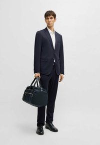 Completo blu navy con una vestibilità su misura, dotato di rever a punta e tasche. Il modello tiene in mano una borsa blu scuro con doppio manico e dettagli in zip.