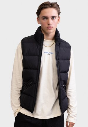 Ung mand iført en sort quiltet vest over en cremefarvet langærmet skjorte med tekst og en guldkæde, stående mod en ensfarvet baggrund.