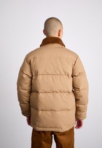 Beige Pufferjacke mit einem braunen Kunstfellkragen, gestepptem Muster und gerippten Bündchen. Das Design hat eine lockere Passform und eine einfache Silhouette.
