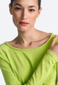 Dlhý rukáv, jasne limetkovo zelená košeľa z mäkkej látky s širokým výstrihom a detailmi s neupraveným okrajom, oblečená modelkou s neutrálnym výrazom.