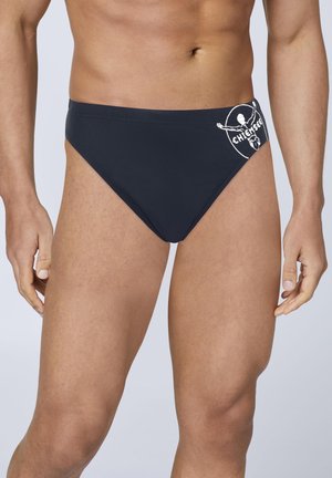 Man wearing black swim briefs with a white logo on the left hip, standing against a plain light background.