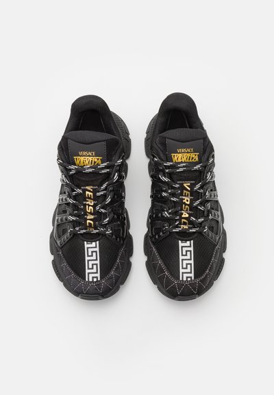 Black athletic shoes with mesh and synthetic materials. Features include reflective accents, bold lacing, and a Greek key pattern on the midsole.