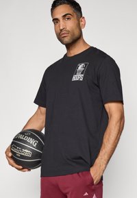 Zwart T-shirt met korte mouwen en een grafische print met een basketbal en de tekst "HOOPS". Model houdt een zwarte Spalding basketbal vast.